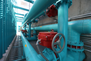 Eine große Gruppe von blauen Rohren und Ventilen in einem Geböude mit einem prominenten roten Ventil in der Mitte, Metallstangen links und einer Wand im Hintergrund.