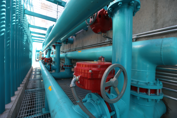 Eine große Gruppe von blauen Rohren und Ventilen in einem Geböude mit einem prominenten roten Ventil in der Mitte, Metallstangen links und einer Wand im Hintergrund.