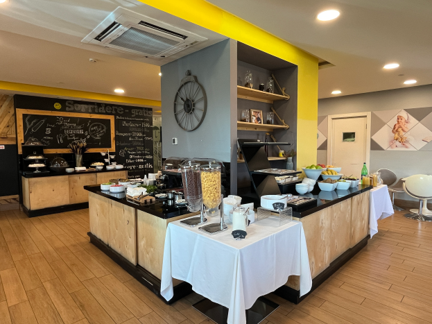 Ein Restaurant-Innenraum mit Tischen und Stühlen, Speisen, einer Tafel mit Text, einer Wanduhr, Deckenleuchten, einer Klimaanlage und einer Tür im Hintergrund, gelegen im Hôtel de Ville in Paris.