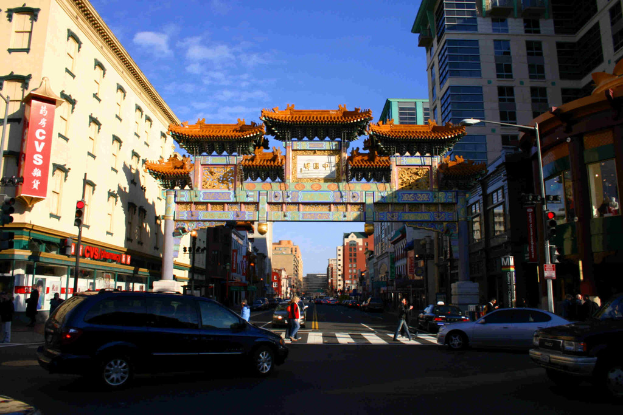 Eine Straße mit fahrenden Fahrzeugen, Fußgänger an einer Zebrastreifenfurt, ein Tor im Hintergrund, Gebäude auf beiden Seiten und eine Ampel.