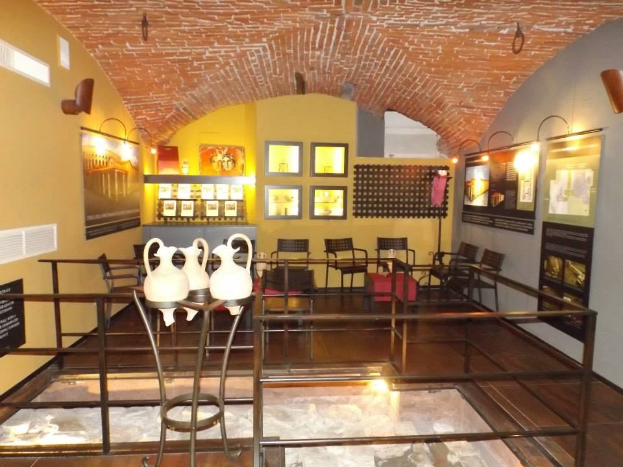 Ein Museumsraum mit mehreren Tischen, Stühlen, Displaytafeln, gerahmten Kunstwerken an den Wänden, Vasen und Deckenbeleuchtung.