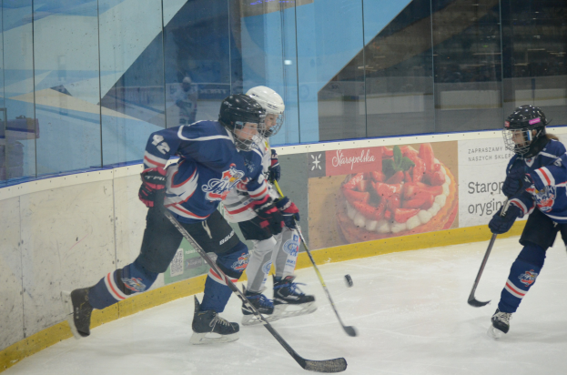 Gruppe junger Menschen, die Eis hockey auf einer Indoor-Eisbahn spielen, mit Helmen, Sportuniformen und Schlittschuhen sowie Hockeystöcken, während im Hintergrund eine Glaswand und ein Plakat zu sehen sind.