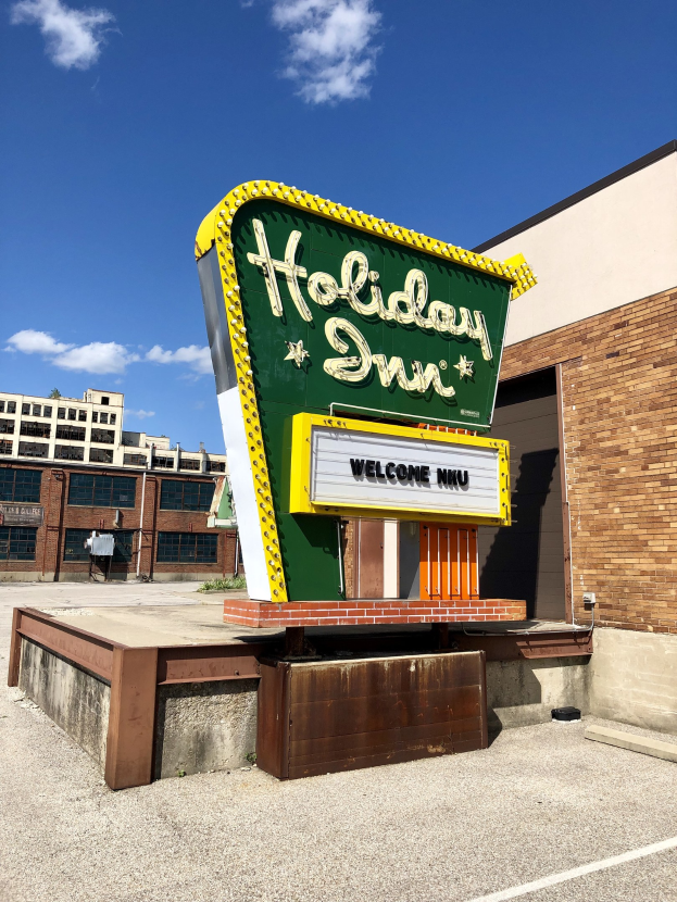 Holiday Inn-Schriftzug an einem Gebäude mit einem Schild auf dem Bürgersteig davor, bewölktem Himmel und sichtbaren Fenstern im Gebäude.