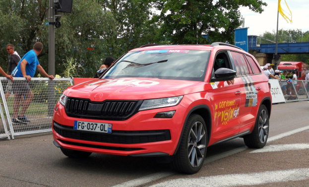 Ein rotes Auto fährt auf einer Straße neben einer Menschenmenge, einige tragen Mützen, mit Maschendrahtzaun, einer Verkehrsampelstange, Bäumen, einer Brücke, einer Fahne an einem Mast und einem bewölkten Himmel im Hintergrund.