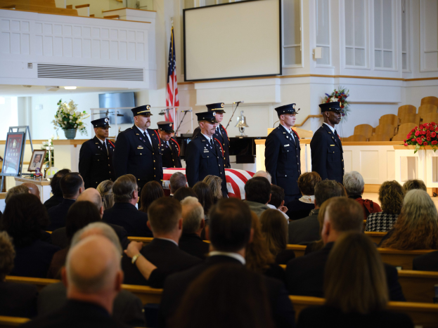 Eine Gruppe uniformierter Personen mit Mützen, die vor sitzenden Trauergästen an einem Grab stehen, mit Blumensträußen, einer Fahne, einem Bildschirm und einer Wand im Hintergrund.