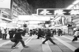 Eine schwarz-weiße Straßenszene mit Menschen auf der Straße, Gebäuden auf beiden Seiten und dem Himmel oben.