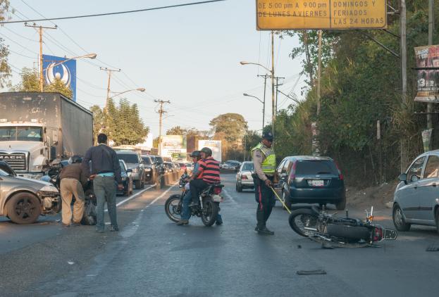 Gruppe von Menschen um ein verunglücktes Motorrad auf dem Seitenstreifen mit mehreren Fahrzeugen, darunter ein Lastwagen, im Hintergrund und Bäumen, Pfosten, Laternen, Schildern und Himmel.