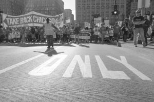Ein Schwarz-Weiß-Bild einer Straßenszene mit Menschen, die gehen, einige mit Schildern und Plakaten, Gebäuden, einem Verkehrssignalmast und Tafeln im Hintergrund.
