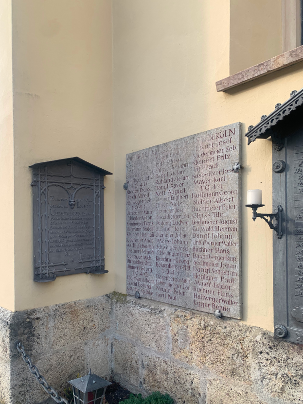 Ein Gebäude mit einer Gedenktafel für Holocaust-Opfer, mit einem Fenster, einer Tür, einem Licht, einer Kette und umgebenden Pflanzen.