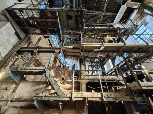 Alter Industriebau mit zentralem Metallgerüst, verrosteten Wänden und zerfallendem Boden, umgeben von Eisenstangen, Treppen und Fenstern.