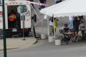 Ein Rettungswagen steht auf der linken Seite einer Straße, während auf der rechten Seite ein Zelt eine Gruppe von Menschen schützt, mit einem Tisch und einem Stuhl in der Nähe.