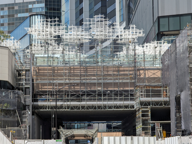 Gro├čes Geb├Ąude mit Gerüst in Bauumgebung mit B├Ąumen links und einer Stra├če unten in einer urbanen Umgebung.