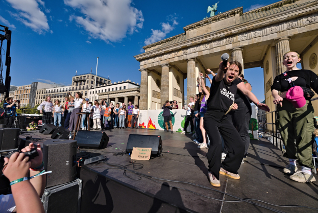 Eine Gruppe von Menschen auf einer Bühne vor dem Brandenburger Tor in Berlin, einige halten Mikrofone oder Kameras, mit Lautsprechern und Equipment im Hintergrund, vor Gebäuden, Bäumen und Himmel.