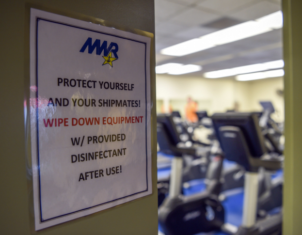 Ein Türschild mit der Aufschrift "Schützen Sie sich und Ihre Schiffskameraden" mit Laufbändern und Deckenleuchten im Hintergrund, mit einem leicht unscharfen, bewegungshaften Effekt.