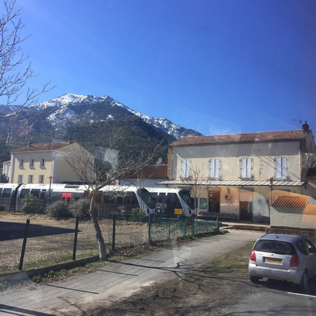 Ein Auto ist auf der linken Seite des Bildes geparkt, mit Zügen, die auf den Schienen vorbeitreiben; Gebäude sind im Hintergrund sichtbar, Bäume sind auf der rechten Seite vor einem Zaun und ein Berg mit Himmel darüber ist im Hintergrund.
