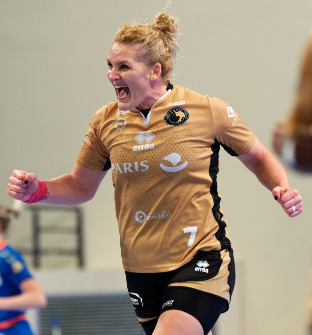 Eine Frau in braunem T-Shirt und schwarzen Shorts rennt auf einem Futsalplatz, feiernd mit triumphaler Miene, während zwei andere Frauen im Hintergrund zu sehen sind.