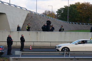 Eine Gruppe von Polizisten steht neben einem Auto auf der Straße, mit Verkehrskegeln, einer Trennwand, Gras, einer Wand, Laternenpfählen, Bäumen und einem bewölkten Himmel im Hintergrund.