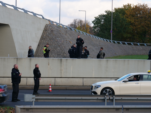 Eine Gruppe von Polizisten steht neben einem Auto auf der Straße, mit Verkehrskegeln, einer Trennwand, Gras, einer Wand, Laternenpfählen, Bäumen und einem bewölkten Himmel im Hintergrund.