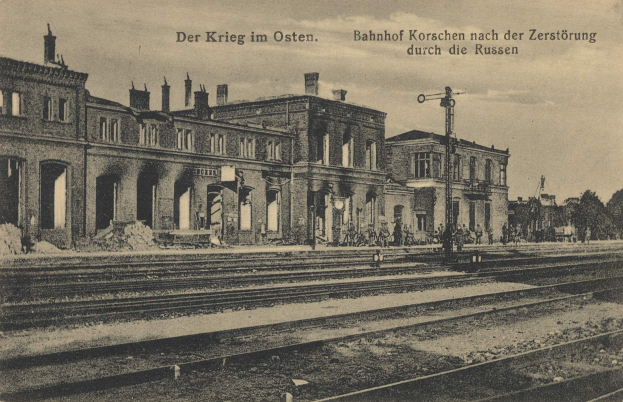 Altes Schwarz-Weiß-Foto des Bahnhofs Korschen in Ostern, Deutschland, mit Bahnschienen, Bahnhofsgebäuden, Menschen, Bäumen und Himmel.