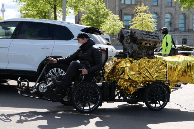 Ein Mann im Rollstuhl mit einem großen Motor auf dem Rücken, umgeben von Fahrzeugen auf einer Straße mit Bäumen, Gebäuden und einem klaren blauen Himmel im Hintergrund, trägt eine schwarze Jacke, eine Mütze und hält ein Objekt.