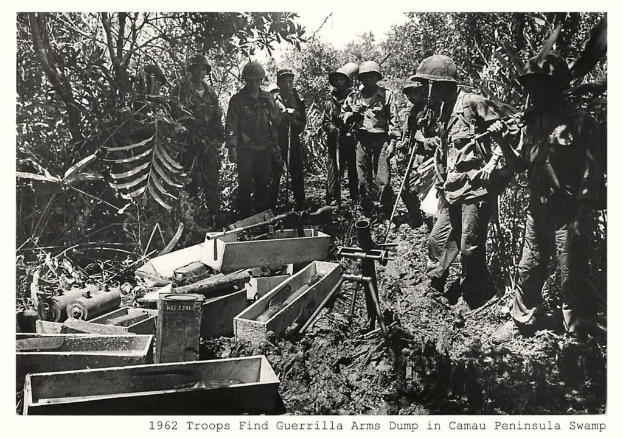 Soldaten in Helmen, Hemden, Hosen und Schuhen stehen vor Holzboxen und Zylindern, mit Bäumen im Hintergrund und einem Himmel darüber und einem Wasserzeichen in der rechten unteren Ecke.