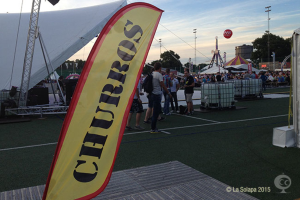 Ein buntes Faschingstreiben mit Zelten, einem zentralen Mast, Menschen, Bäumen, Strommasten, Straßenbahnen auf der Straße und einem Eisenständer links.