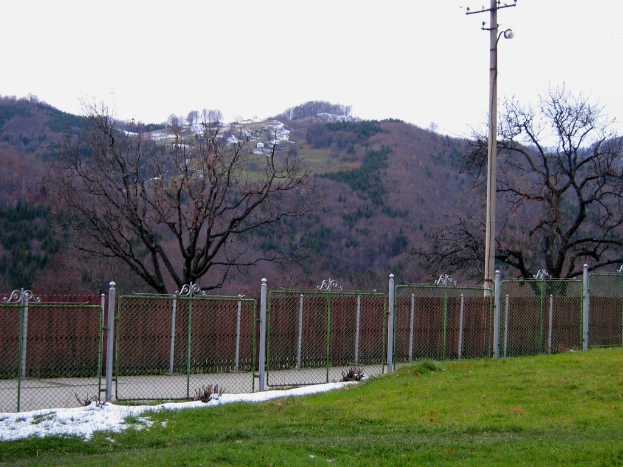 Eine grüne Landschaft mit einem Zaun und einem Pfahl im Vordergrund, Bäumen im Hintergrund und ein paar Häusern auf einem Hügel.