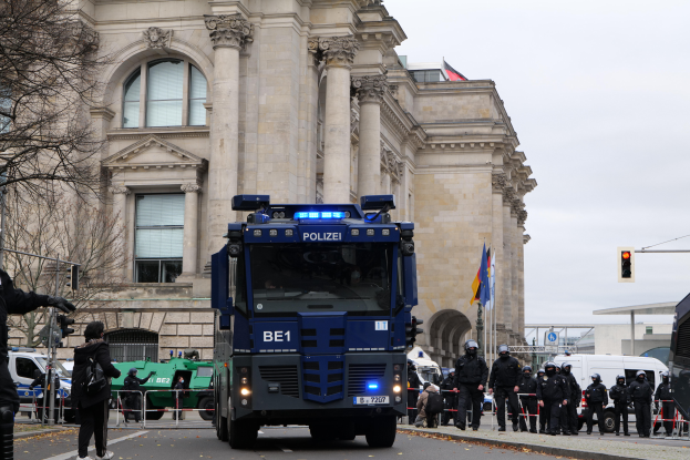 Eine Gruppe von Polizisten steht vor einem großen Gebäude mit Fenstern, Säulen und Bögen, mit Fahrzeugen auf der Straße, einer Person mit einer Kamera auf der linken Seite und einem klaren blauen Himmel mit Bäumen, Ampeln und Flaggen im Hintergrund.