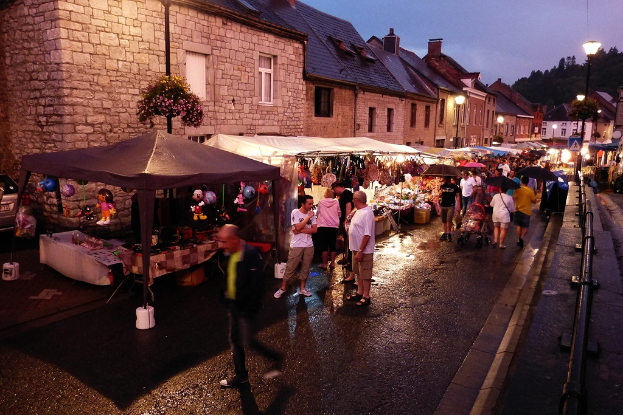 Eine Gruppe von Menschen, die auf einer Straße mit Geschäften, Schirmen, hängenden Spielzeugen, Taschen, Gebäuden mit Fenstern, Bäumen, Straßeleuchten und Barrieren spazieren gehen.