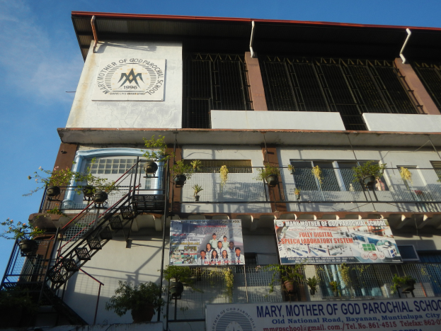 Das Gebäude der Mariä-Mutter-Gottes-Grundschule mit Fenstern, Geländern, Treppen, Blumentöpfen, Bannern und einem bewölkten Himmel im Hintergrund.