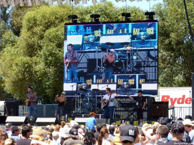 Eine Band performs auf der Bühne mit verschiedenen Musikinstrumenten und Mikrofonen, ein digitaler Bildschirm hinter ihnen zeigt ihre Performance und ein Publikum schaut ihnen zu.