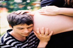 Ein Mann und eine Frau stehen nebeneinander, der Mann berütht zart den Bauch der Frau, mit einem unscharfen Hintergrund, der das Paar und ihr ungeborenes Kind betont.