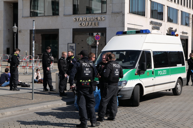 Eine Gruppe von Polizeibeamten steht vor einem Starbucks-Café mit einem Van auf der rechten Seite und einigen Menschen auf der linken Seite. Im Hintergrund gibt es ein Gebäude, ein Schild, einen Lichtmast und eine Absperrung.