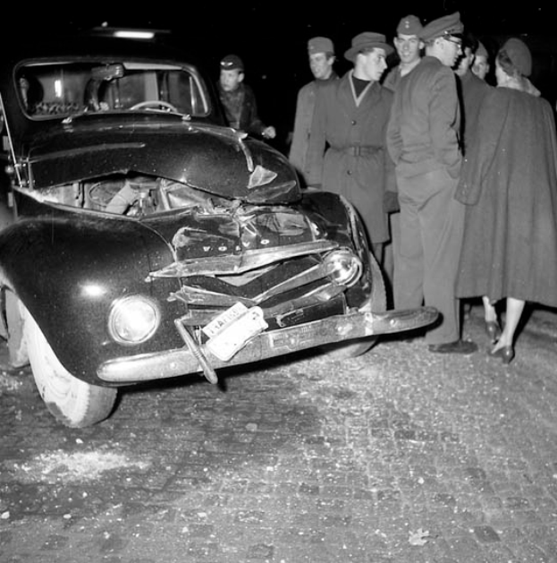 Ein Schwarz-Weiß-Bild eines beschädigten Autos am Straßenrand, mit einer Gruppe besorgter Menschen drumherum.