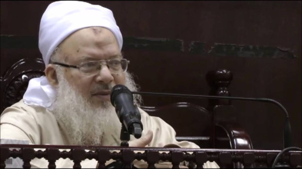 Asaduddin Owaisi, ein Mann mit einem weißen Bart und Turban, spricht in ein Mikrofon und trägt ein cremefarbenes Gewand und eine Brille, vor einem hölzernen Geländer gegen einen dunklen Hintergrund.