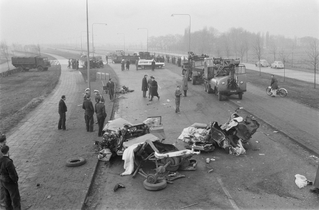 Schwarz-weiß-Szene eines Autounfalls am Straßenrand mit mehreren Fahrzeugen, darunter ein Landfahrzeug, und einer Gruppe von Menschen, die darum herumstehen, mit Laternenpfählen, Bäumen und dem Himmel im Hintergrund.