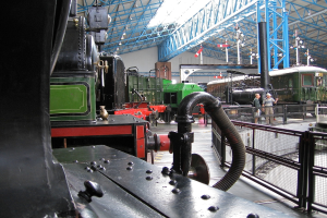 Lokomotiven fahren mit Eisenstangen an der Spitze, ein Zaun auf der rechten Seite, ein Zug auf dem Bahngleis neben dem Zaun und zwei Personen in der Mitte nahe dem Zaun.