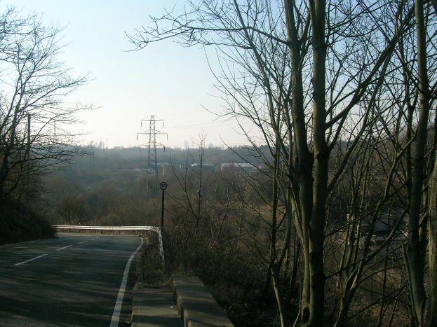Eine Straße mit einer Barriere, Übertragungsmast, Stromleitungen und Bäumen unter einem klaren Himmel.