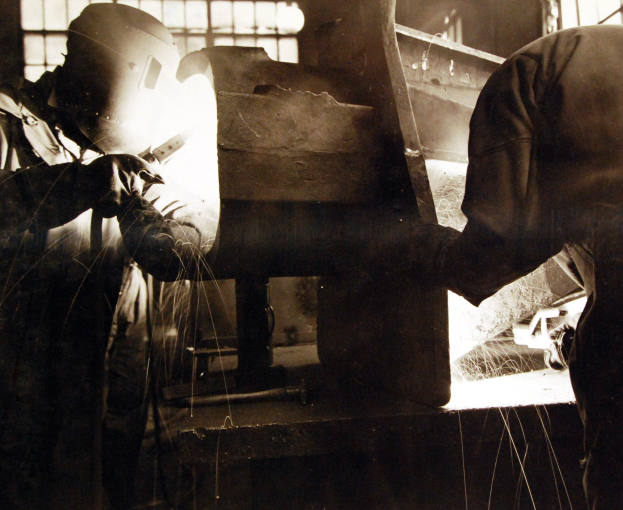 Zwei Männer in Schutzausrüstung, einschließlich Helmen und Handschuhen, schweißen ein Metallteil mit einer Schweißmaschine in einer Fabrik mit einer Fensterwand im Hintergrund, dargestellt in Schwarz-Weiß.