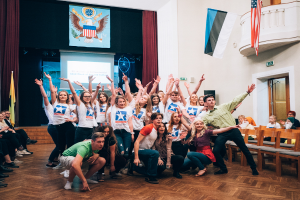 Eine Gruppe von Menschen, die vor einer sitzenden und stehenden Menge posiert, mit einem Bildschirm, Vorhängen, Fahnen, einem Schild, einer Tür, einem Geländer und einer Decke mit Deckenleuchten im Hintergrund, wahrscheinlich auf einem nationalen Jugendkongress.