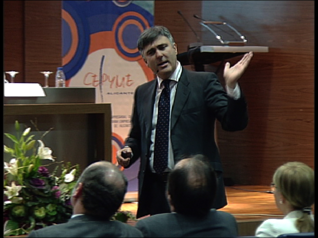 Zwei Männer und eine Frau sitzen auf Stühlen, während ein Mann in einem schwarzen Blazer an einem Podium mit Mikrofonen steht und in ein Handsender spricht, mit einem Blumentopf oder einem Blumenstrauß in der Nähe und einem Tisch mit einem Buch, einer Wasserflasche und anderen Gegenständen daneben, vor einer braunen Wand und einer bunten Tafel mit Text im Hintergrund, wahrscheinlich in einem Konferenzsaal.