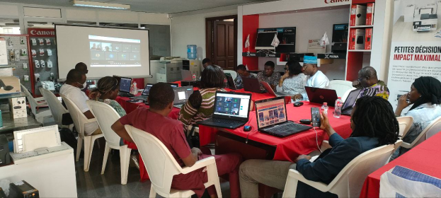 Eine Gruppe von Menschen sitzt um einen Tisch mit Laptops, Mäusen und Flaschen, nimmt an einem digitalen Marketing-Workshop teil, mit einem Banner, Display-Bildschirm und elektronischen Geräten im Hintergrund.