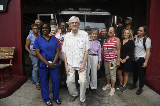 Gruppe von Freiwilligen vor einem Feuerwehrauto, mit einer Bank auf der linken Seite und einer Wand im Hintergrund.