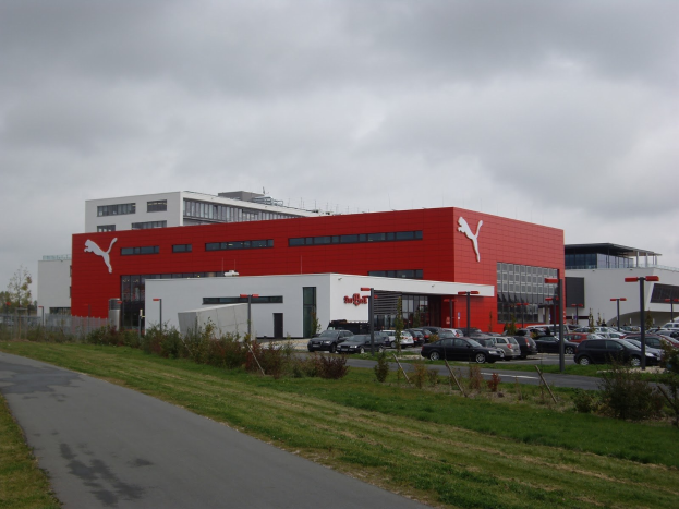 Fabrikgebäude von Puma in Deutschland mit parkenden und fahrenden Fahrzeugen, Straßeninfrastruktur, Grünflächen und einem bewölkten Himmel.