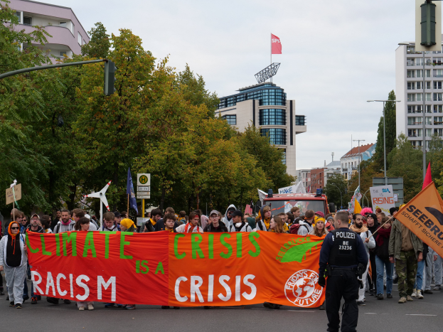Eine Gruppe von Menschen marschiert eine von Bäumen gesäumte Straße entlang und hält ein Schild mit der Aufschrift "Klima-Krise ist eine Krise" hoch, mit Gebäuden und einem klaren blauen Himmel im Hintergrund.