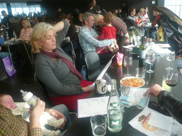 Ein Innenblick in ein belebtes Restaurant mit Menschen an Tischen, die mit Gläsern, Tellern mit Essen, Getränkeflaschen, Speisekarten und anderen Gegenständen gedeckt sind.