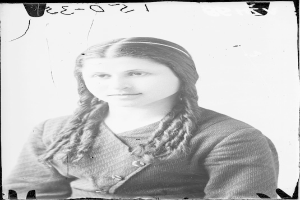 Ein Schwarz-Weiß-Porträt von Julia Margaret Cameron, eine Frau mit langen Zöpfen, die ein weißes Kleid und einen gemusterten Schal trägt und einen sanften Ausdruck hat und ihre Hände vor der Brust verschränkt.