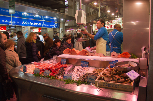 Ein belebtes Marktbild mit Menschen, die gehen und Fisch schneiden, Fisch und Bretter unten und Läden, Bretter, Pfähle und Lichter im Hintergrund.