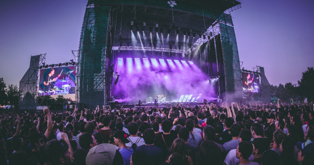 Eine große Menschenmenge auf einem Musikfestival mit einer Bühne in der Mitte, auf der Musiker Instrumente spielen, beleuchtet von Lichtern und umgeben von Bildschirmen, mit einem Gebäude, Bäumen und einem klaren blauen Himmel im Hintergrund.