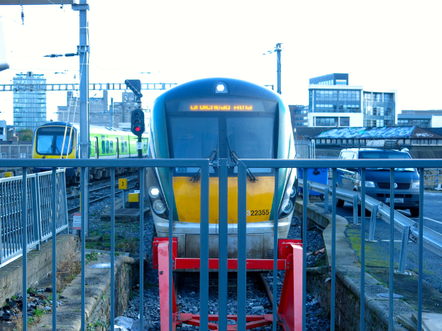 Zwei Züge auf Schienen mit einem Zaun im Vordergrund, Masten mit einem mit einer Signalleuchte, ein Auto auf der Straße rechts, Gebäude im Hintergrund und Himmel darüber.